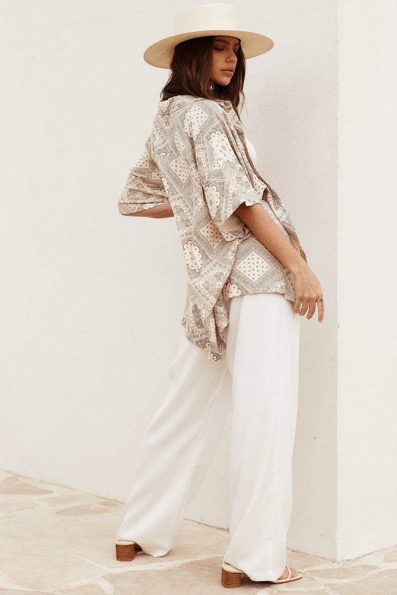 Model in white wide leg trousers styled with paisley blouse, straw hat, and heeled sandals.