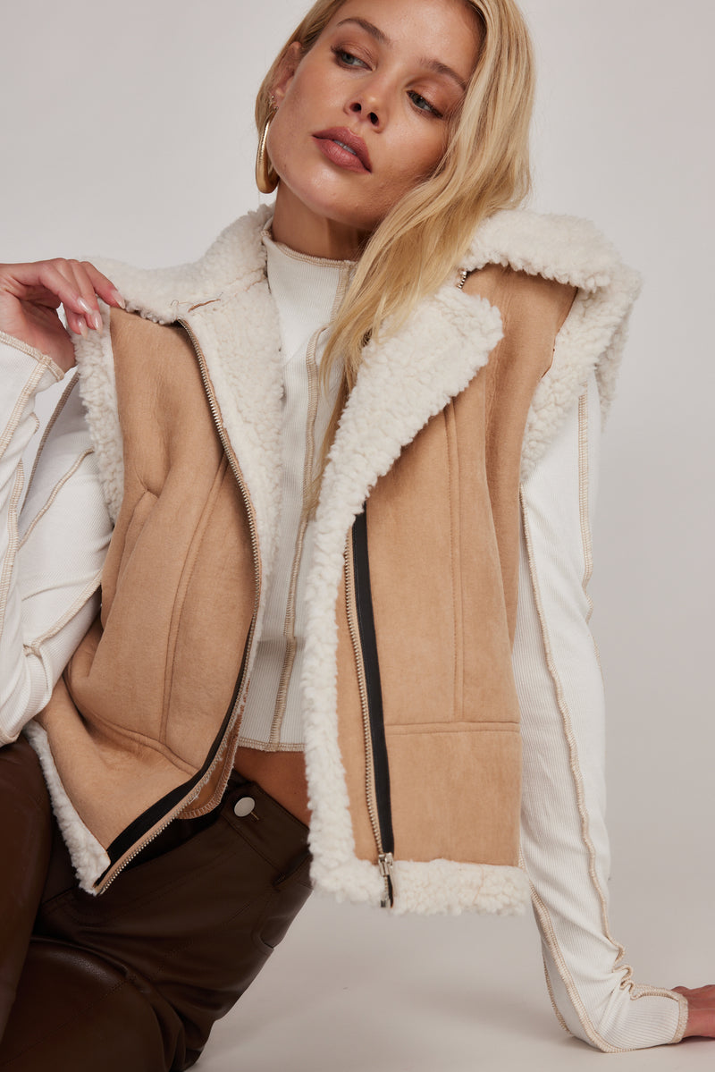 Beige sherpa lined vest worn open, showing faux fur collar and zip front.